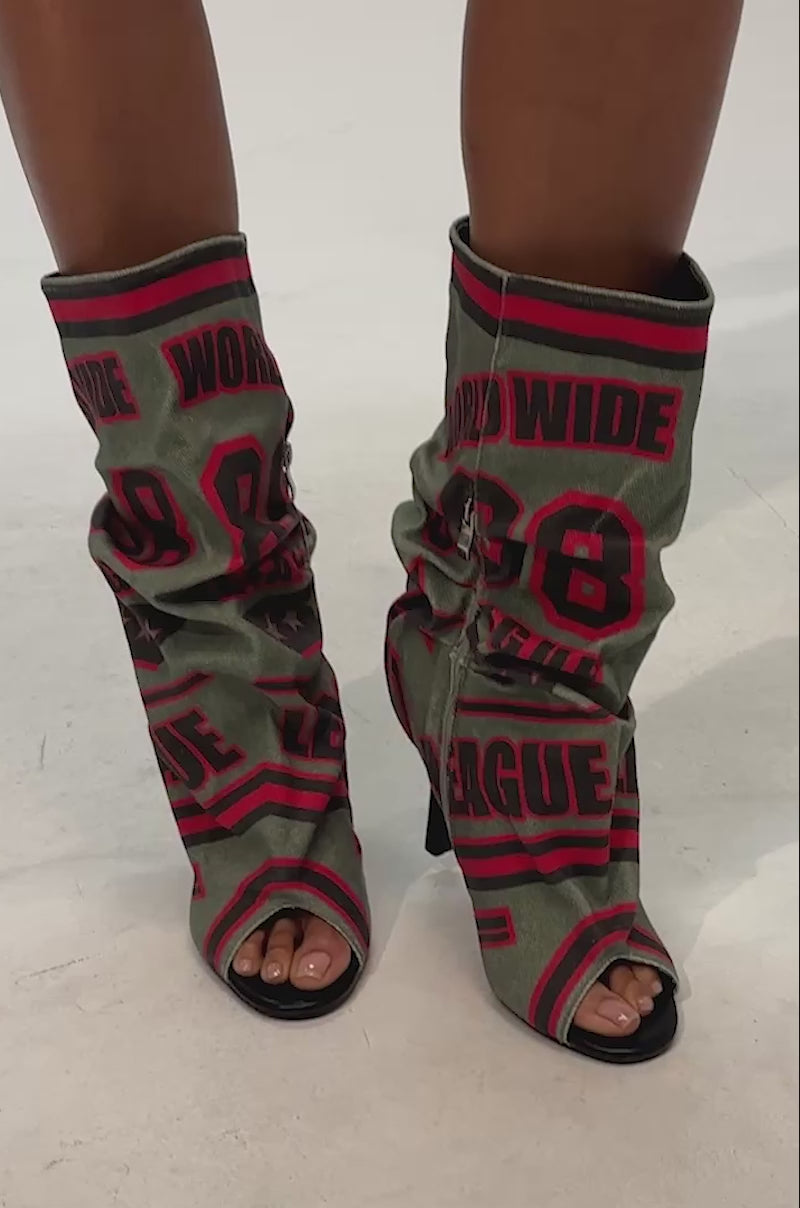 Video of woman modeling the AKIRA Gold Big Time Green Multi Canvas Printed Slouch Open Toe Bootie, featuring olive green tones with bold red and black graphic designs—such as numbers and phrases like "WORLD WIDE" and "LEAGUE"