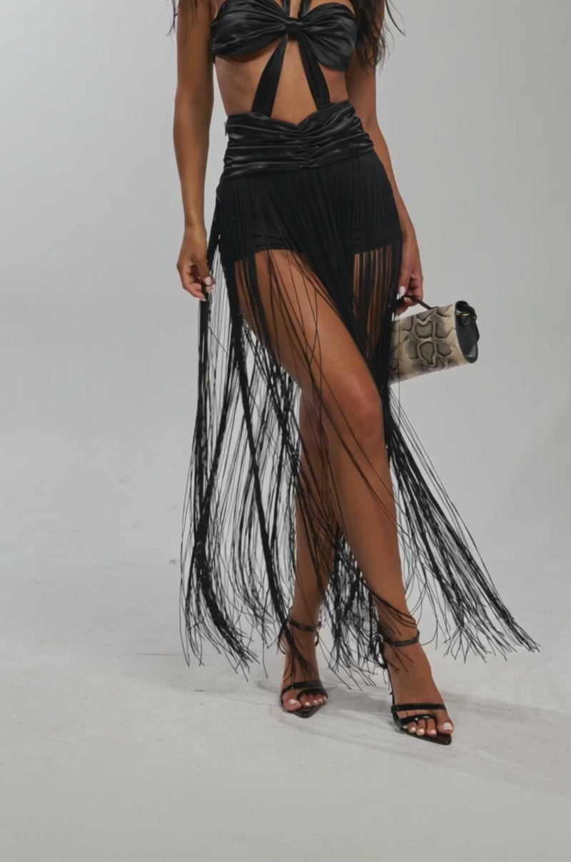Video of woman modeling the TWIST IT UP FRINGE MAXI DRESS, featuring black cutouts and long fringe. Paired with strappy black heels and a chic handbag, this outfit is perfect for formal events. The background is plain and light-colored.