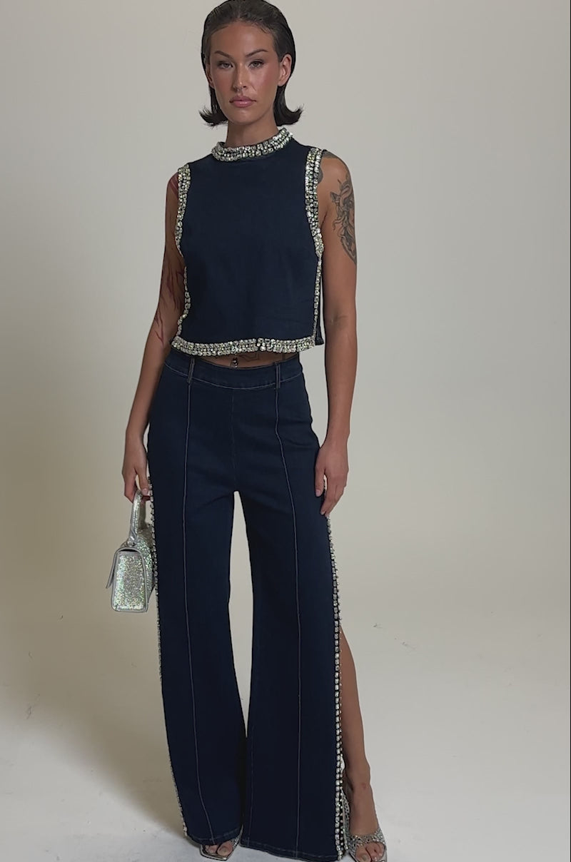 A video of a woman in the POPPIN OUT RHINESTONE DENIM PANT IN DARK WASH DENIM, paired with a sleeveless matching top, silver heels, a sparkly silver handbag, and short slicked-back hair. Silver embellishments trim her chic look.