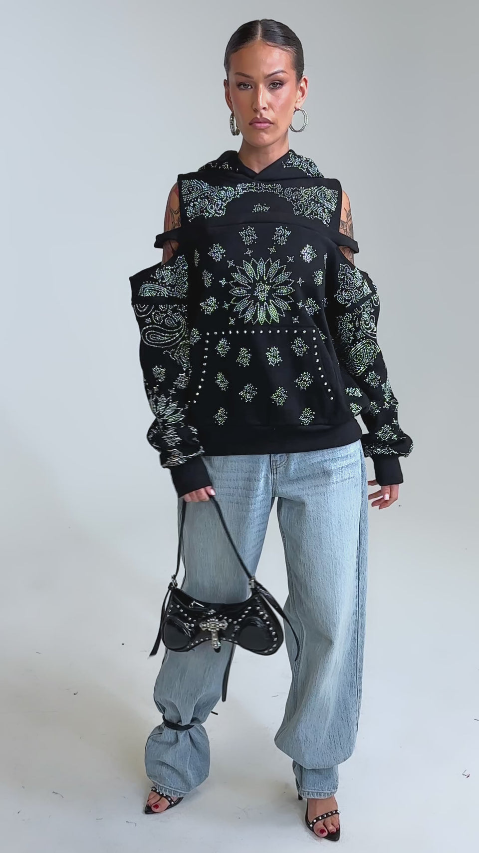 Video of woman modeling the FEELING FREE SWEATSHIRT—a black hoodie with cut-out shoulders and silver details—pairs it with light blue jeans, hoop earrings, slicked-back hair, and visible arm tattoos, standing against a neutral background.