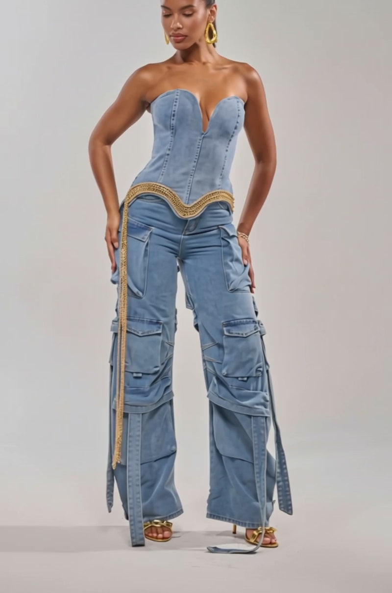 Video of woman modeling the SHOW IT OFF DENIM CORSET, a strapless sweetheart denim corset with gold chain fringe, paired with matching wide-leg cargo jeans, gold jewelry, and heeled sandals, standing against a plain background.
