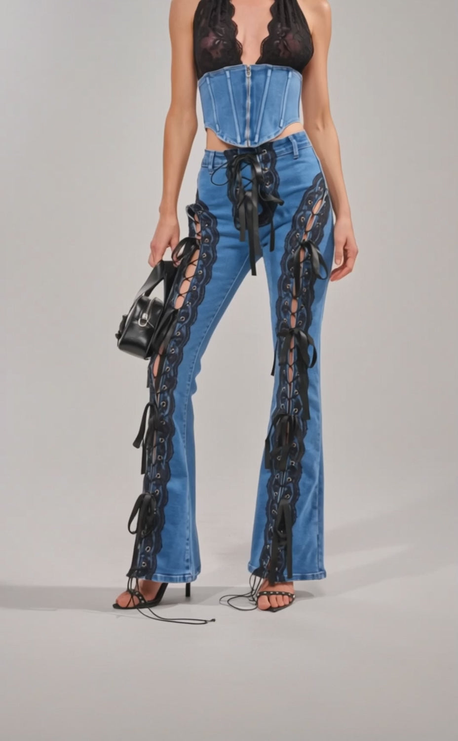 Video of woman modeling the LACED UP DENIM flared pants, featuring black lace-up details, side cutouts, and multiple black ribbon ties for an adjustable bungee cord effect, holding a black handbag.
