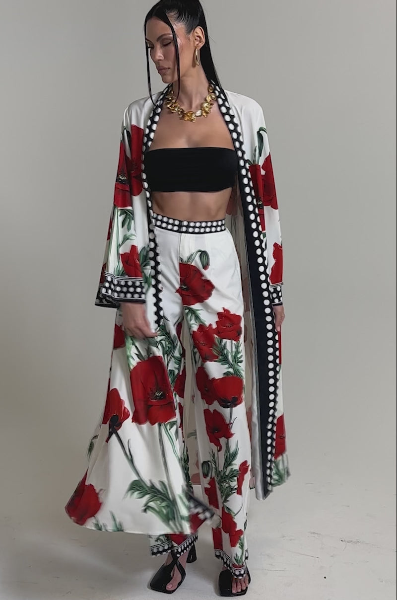 A video of a woman wearing the BELLADONNA CHIFFON DUSTER—a white longline kimono with large red floral prints—paired with matching wide-leg pants, a black bandeau top, black sandals, and a gold necklace against a plain backdrop.