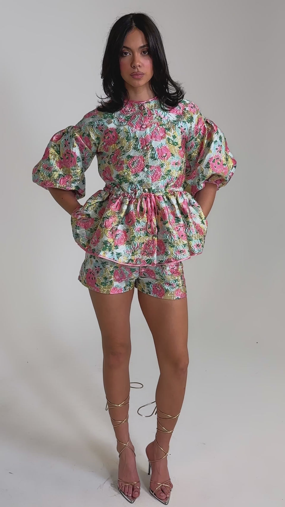 A video of a woman in the TEA TIME FLORAL BROCADE SHORT set, featuring vintage-inspired floral shorts with puffed sleeves and a peplum waist. 