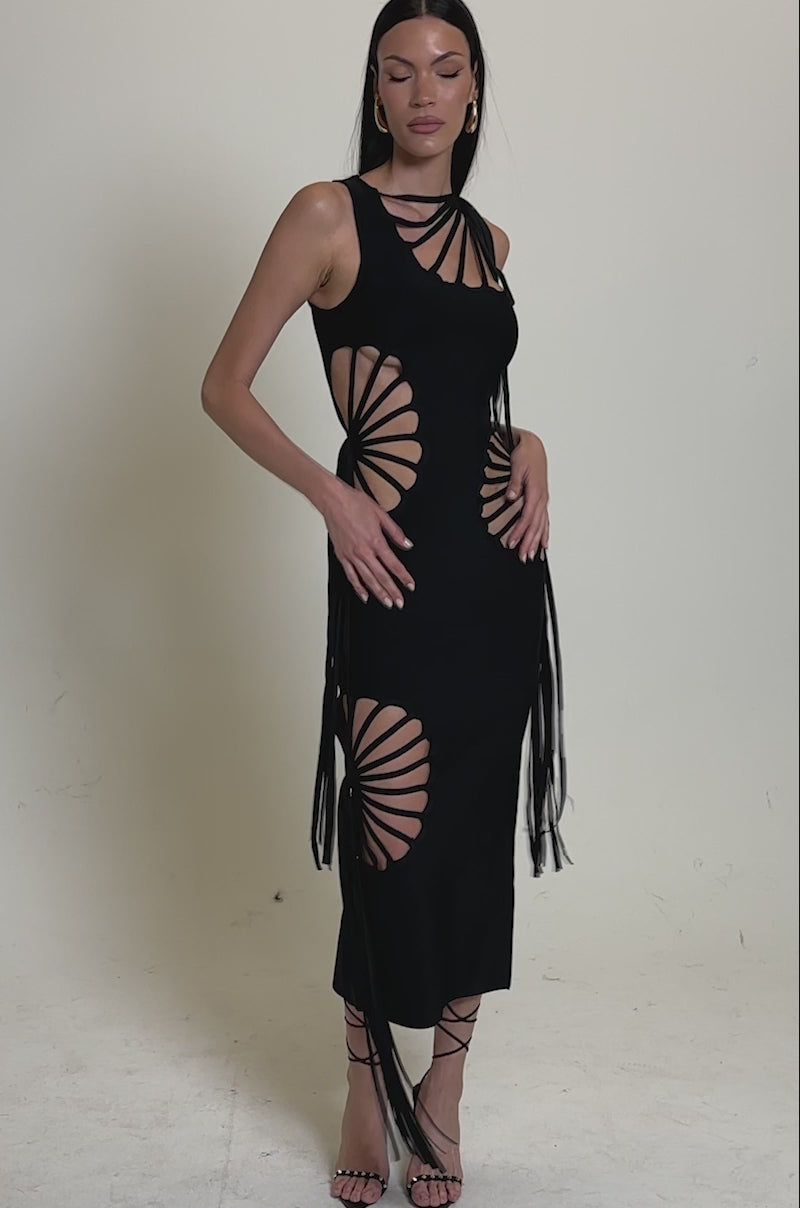 A video of a woman wearing the BROOKE CUTOUT MIDI DRESS, a sleeveless black bodycon midi featuring dramatic cut-outs, radiating strap details on the sides and chest, long dangling ties, and strappy high-heeled sandals.