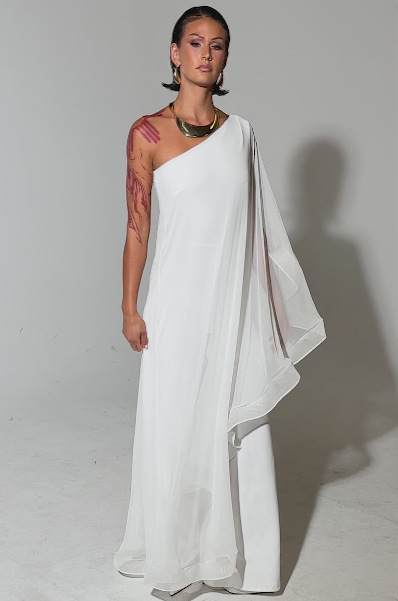A video of a woman in the SANDO MESH JUMPSUIT IN WHITE, a sheer one-shoulder style with wide legs. She wears gold jewelry, has short dark hair and red markings on her arms, set against a plain light gray background.