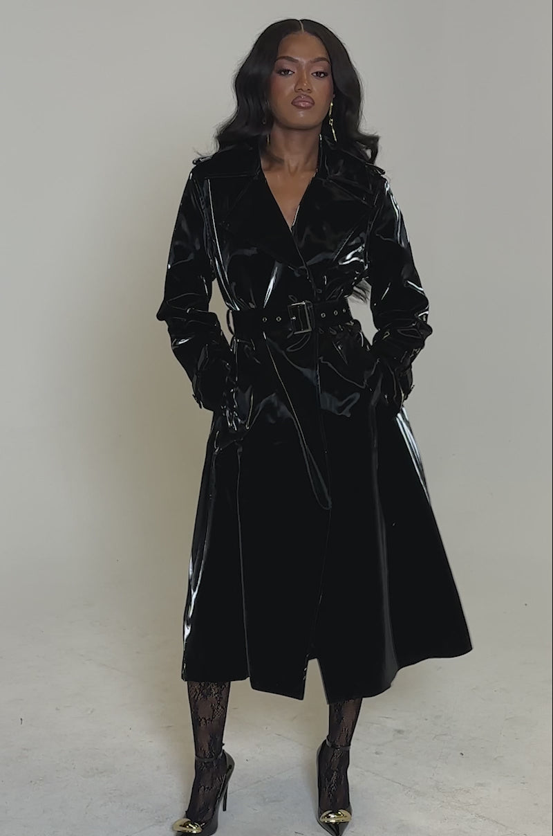 A video of a woman wearing the ONYX LIQUID VELVET TRENCH, paired with black lace tights and black pointed heels with gold tips. Her long wavy hair complements the look as she poses against a plain light background.