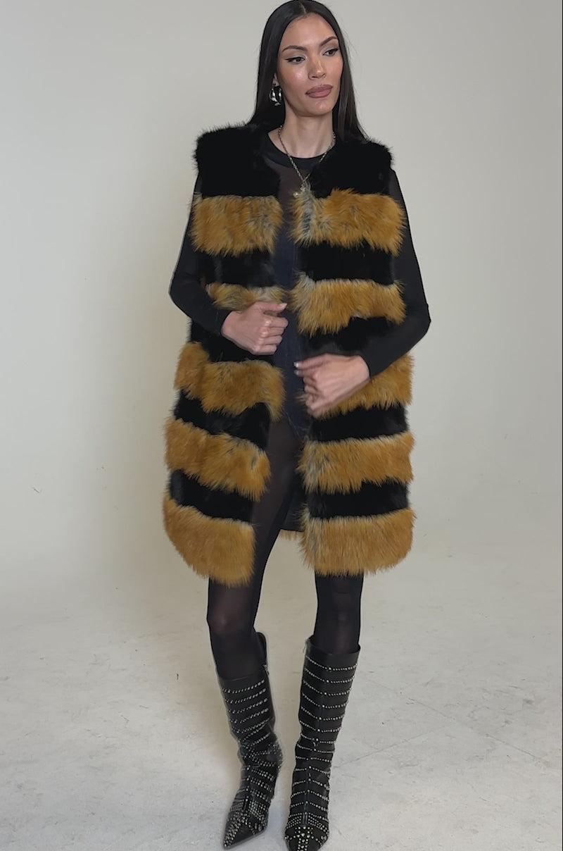 Video of woman modeling the IN ANOTHER LIFE PANELED FUR VEST—a sleeveless, longline faux fur vest with bold black and orange stripes—over black clothes, standing against a plain light background.