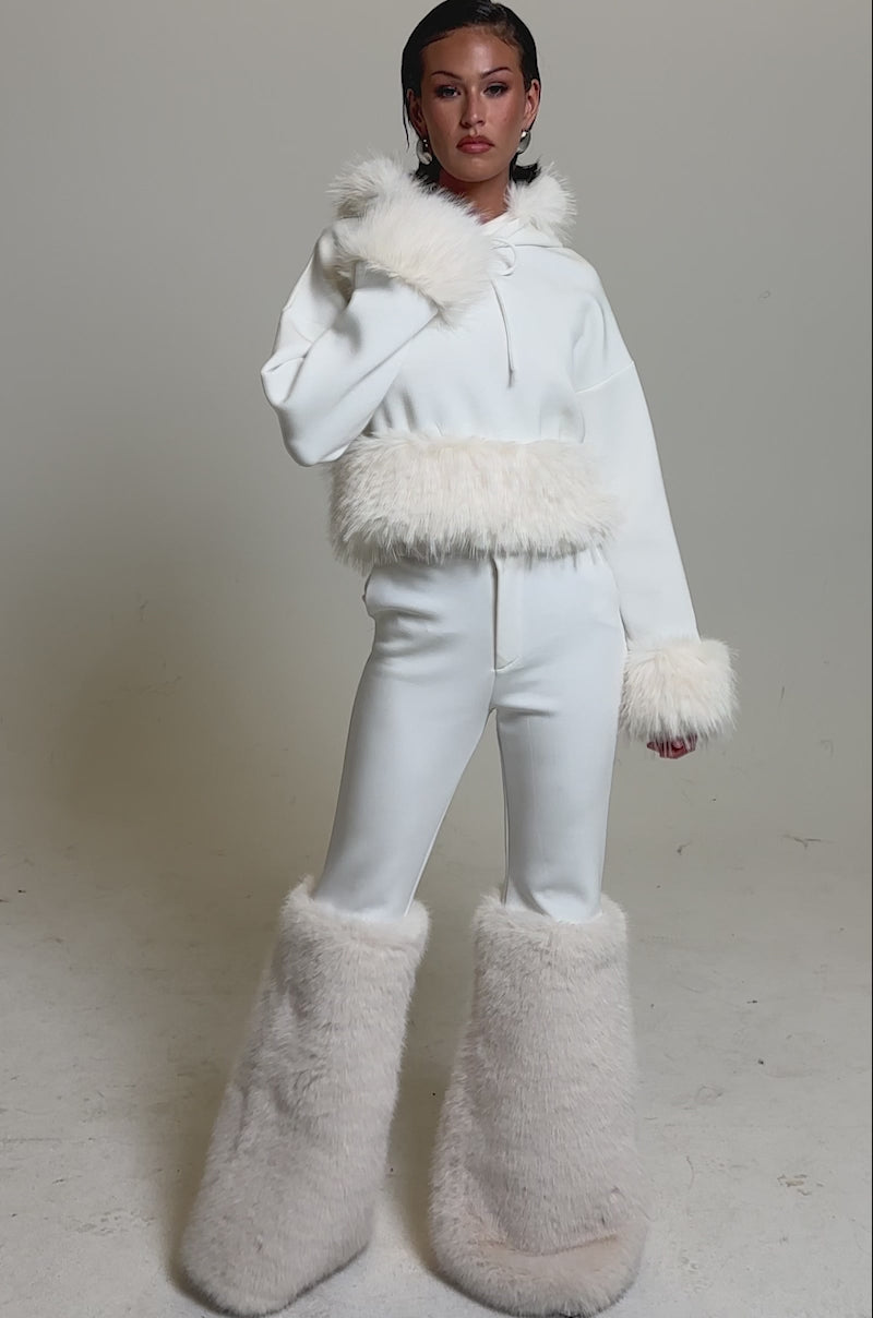 A video of a woman wearing the SNOW WHITE FUR TRIM SWEATSHIRT with fluffy faux fur on the hood, cuffs, and waist, paired with oversized fur boots.