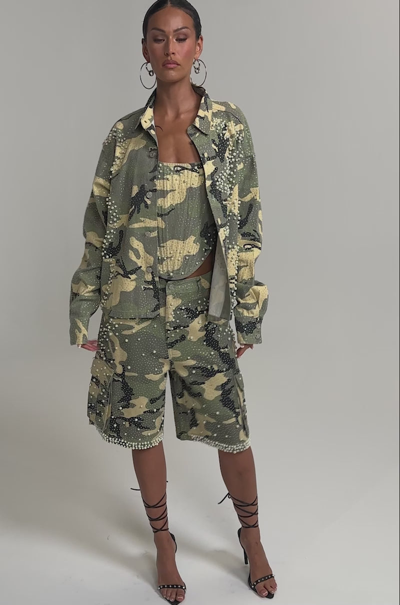 Video of woman modeling the EMBELLISHED CAMO BUTTON DOWN with matching pants, hands in pockets. She wears hoop earrings and a sleek hairstyle, exuding a modern street-luxe vibe against a plain backdrop.