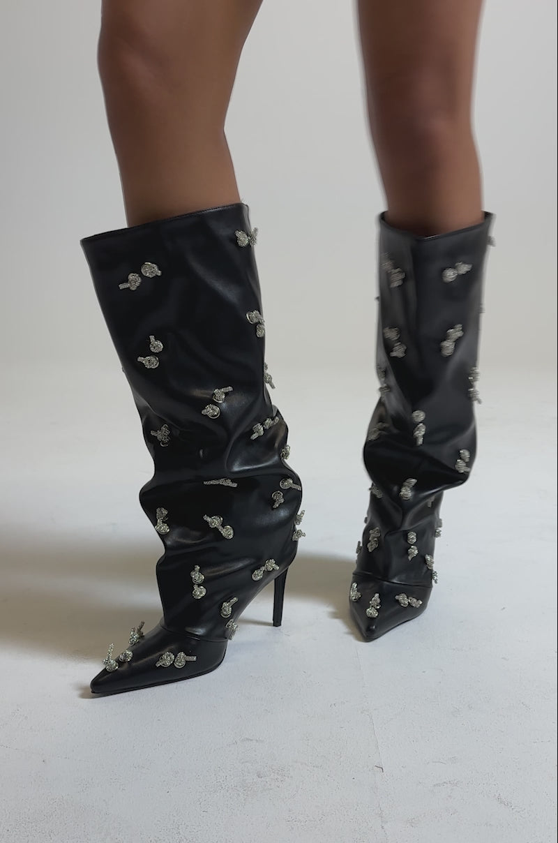 A video of a person wearing the AZALEA WANG COLEMAN STILETTO SILVER HARDWARE BLACK PU BOOT, featuring knee-high black faux leather, pointed toes, and sparkling rhinestone embellishments.
