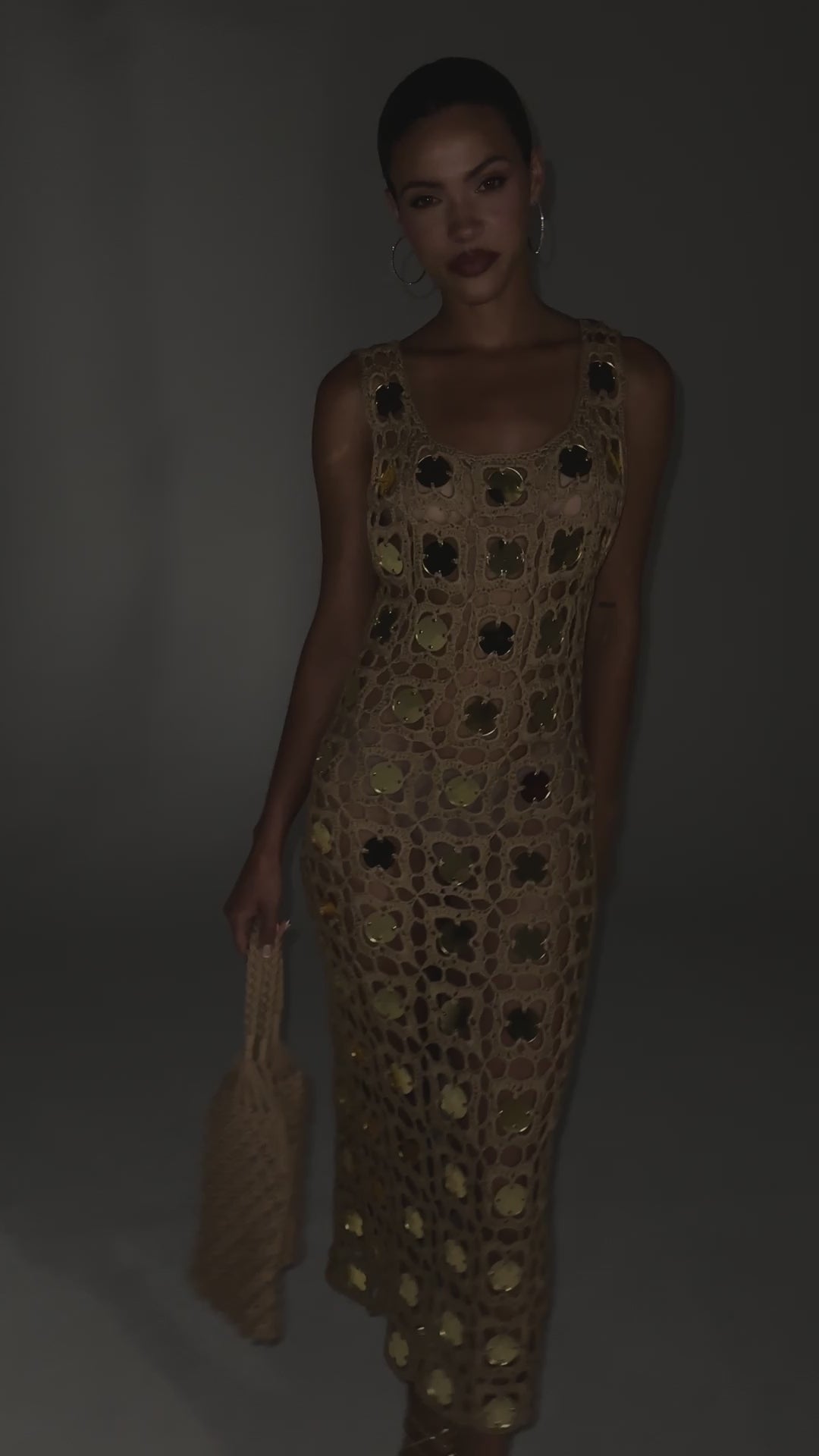 A video of a model wearing the HOT GIRL AGENDA SEQUIN CROCHET MIDI DRESS. A beachy-glam crochet midi dress designed to turn every vacation moment into a statement. Featuring an open-knit crochet construction with shimmering reflective gold disc embellishments throughout, a sleeveless body-skimming silhouette, and a sexy low-cut back.