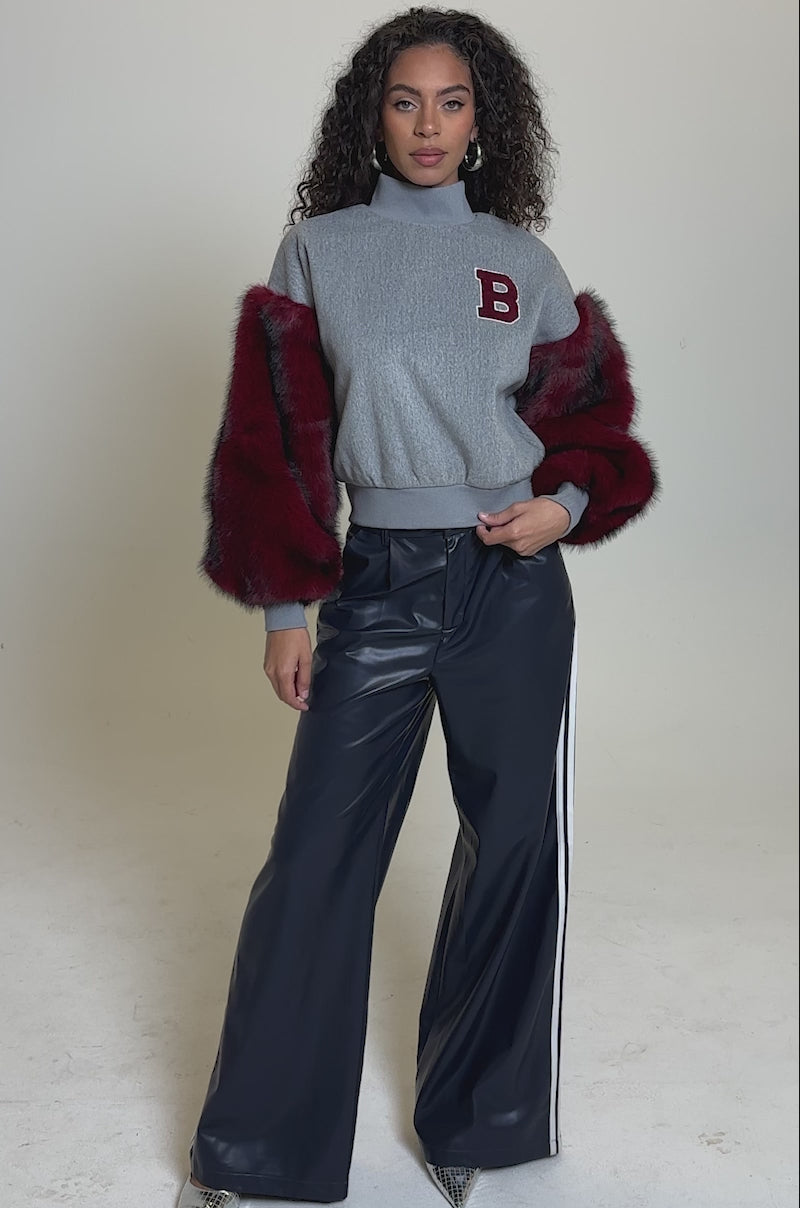 Video of woman modeling the NEW EAST SIDE FAUX FUR SLEEVE SWEATSHIRT with red faux fur sleeves and a red "B" patch, hands in pockets, paired with shiny dark blue pants