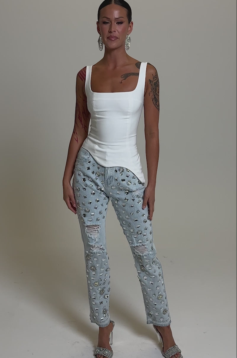 A video of a woman in a fitted white sleeveless top, DAMN GIRL EMBELLISHED SKINNY DENIM jeans, and silver high-heeled sandals.