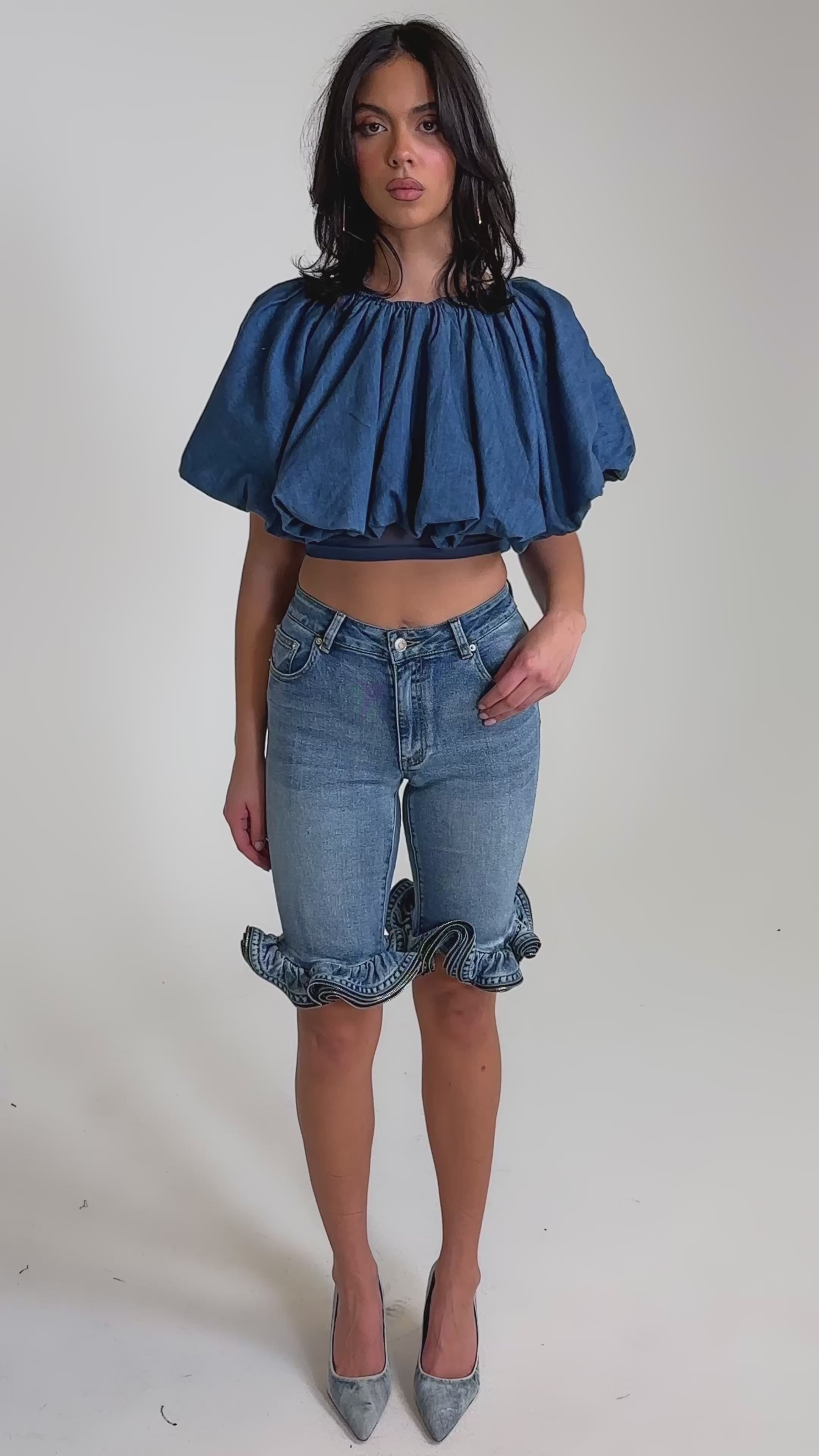 A video of a woman in the NEVER GOING TO STOP BLOUSE IN MEDIUM BLUE DENIM, paired with fitted denim shorts and gray high heels against a plain light background.