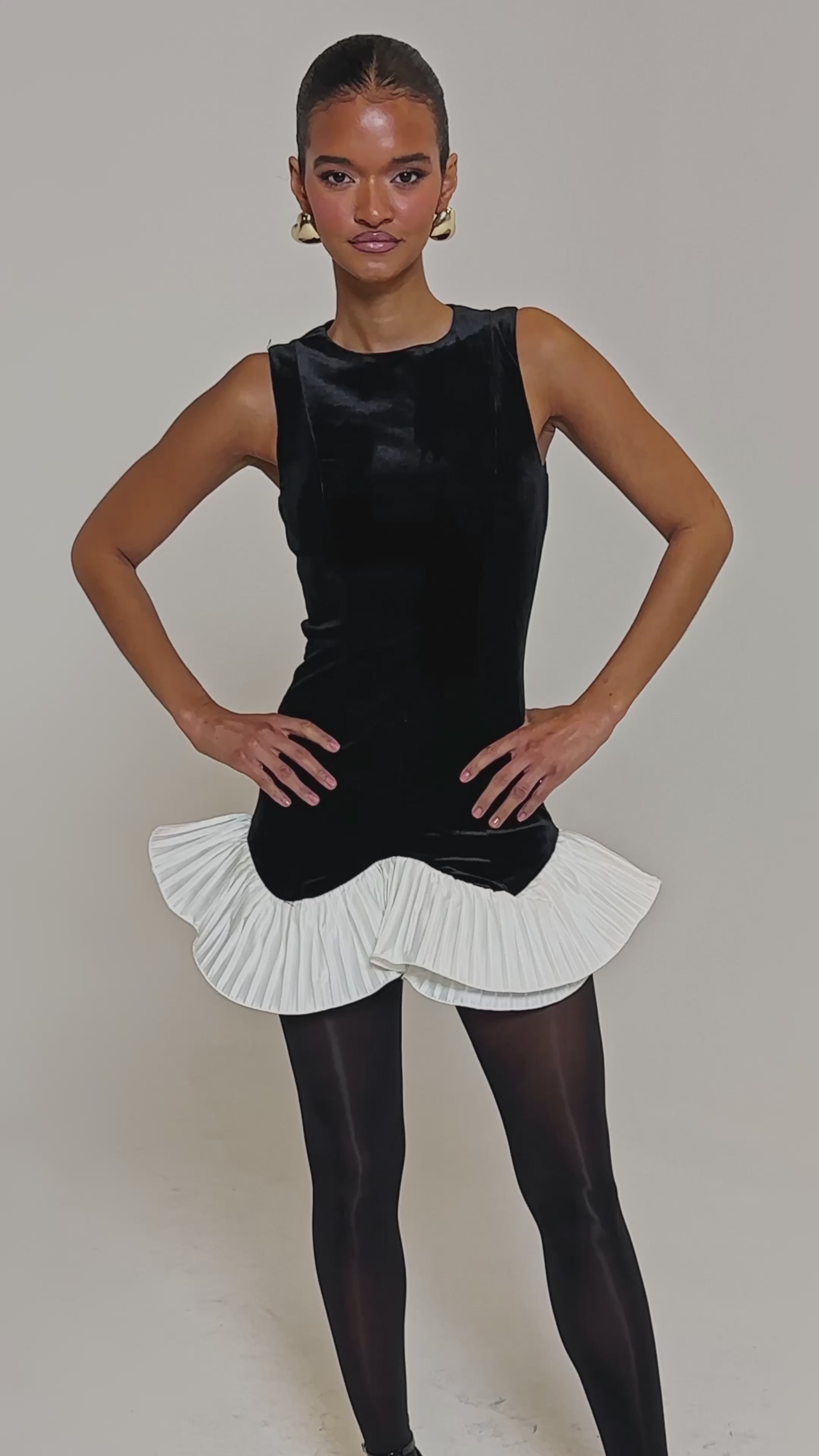 Video of woman modeling the BLACK SWAN VELVET MINI DRESS, featuring a white pleated ruffle hem, styled with black tights, gold-accented black heels, slicked-back hair, and gold hoop earrings for a chic cocktail look.