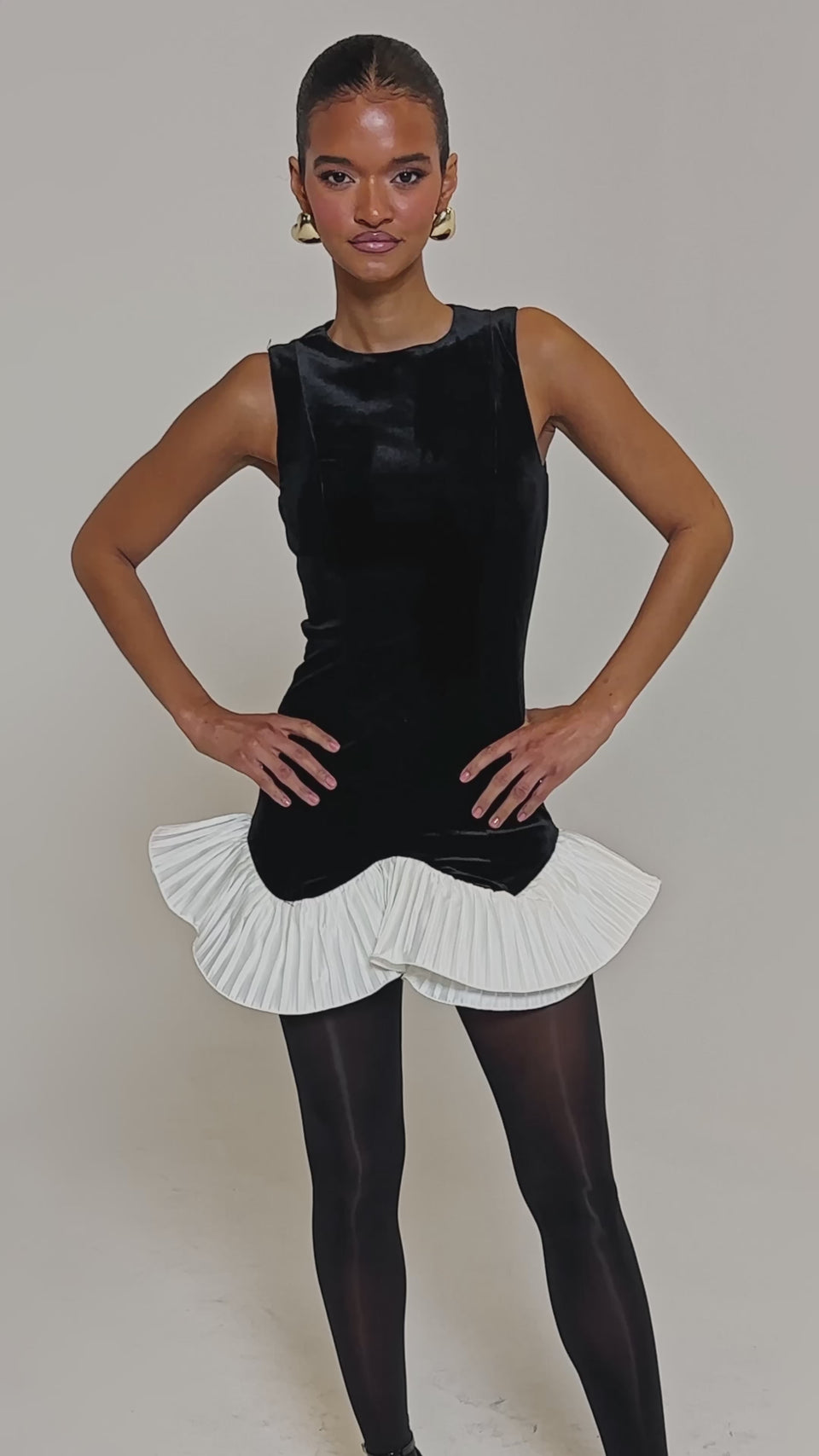 Video of woman modeling the BLACK SWAN VELVET MINI DRESS, featuring a white pleated ruffle hem, styled with black tights, gold-accented black heels, slicked-back hair, and gold hoop earrings for a chic cocktail look.