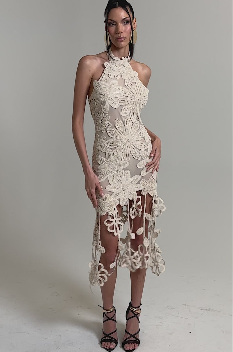 A video of a woman in the CELINE FLORAL MIDI DRESS—an ivory crochet lace midi with bold floral patterns and long fringe details, styled with black strappy heels for a chic look.