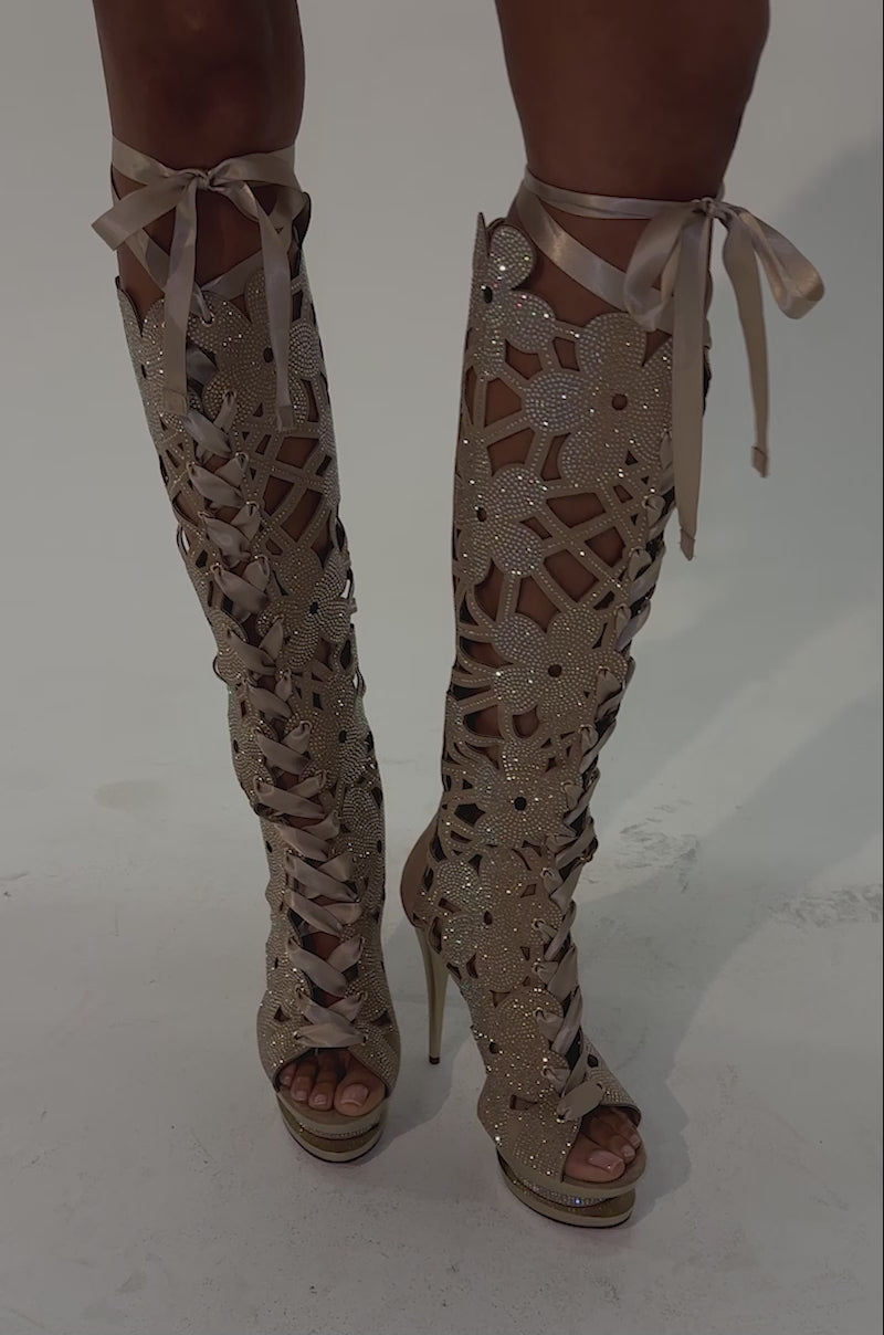Video of a woman modeling the AKIRA Gold Twinkle Rhinestone Embellished Gladiator Sandal—tall, open-toe heels featuring elegant floral cut-out details, sparkling rhinestone embellishments, and ribbon laces.