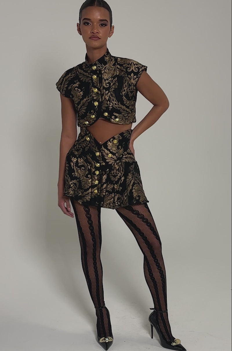 Video of woman modeling the KAYLA BROCADE MINI SKIRT with a matching crop top, styled with sheer black patterned tights and black pointed heels featuring gold tips.
