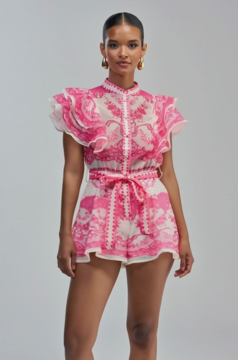 Video of a woman modeling the PRETTY IN PINK PATTERNED ROMPER—a short, pink and white romper featuring ruffle detailing on the sleeves and a tied waist.