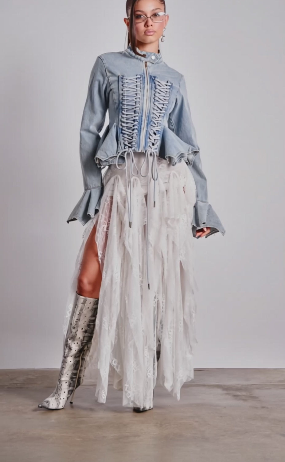 A video of a woman wearing the DOUBLE DARE YOU PEPLUM DENIM JACKET, styled with a flowing white lace skirt, silver knee-high boots, and clear glasses.