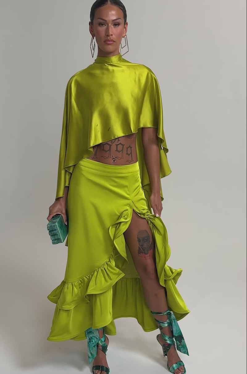 Video of woman modeling the LADY LUXE RUCHED MIDI SKIRT WITH HIGH SLIT in lime green, paired with a matching cropped top, teal clutch, teal ribbon-tied sandals, geometric earrings, and a sleek tied-back hairstyle.