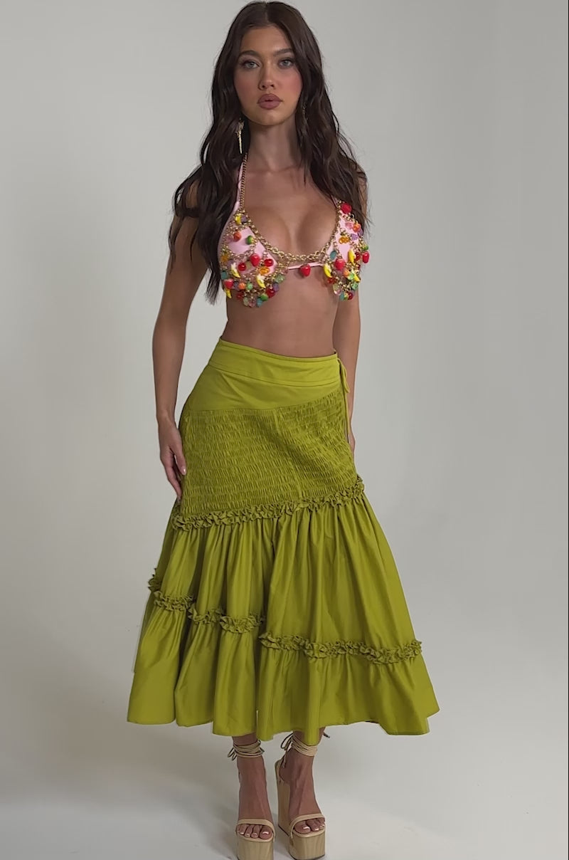 A video of a woman wearing a colorful floral bikini top, a high-waisted yellow tiered skirt, beige platform sandals with ankle ties, and the TASTE ME FRUITY BODY CHAIN.