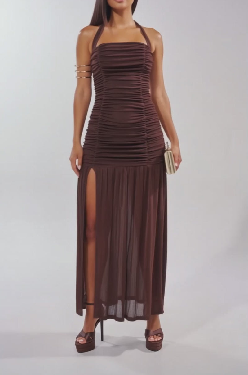 A video of a woman in the NOTHING SUBTLE ABOUT IT RUCHED MAXI DRESS IN BROWN with a high slit. She pairs it with a light clutch, brown platform heels, and a gold arm cuff.