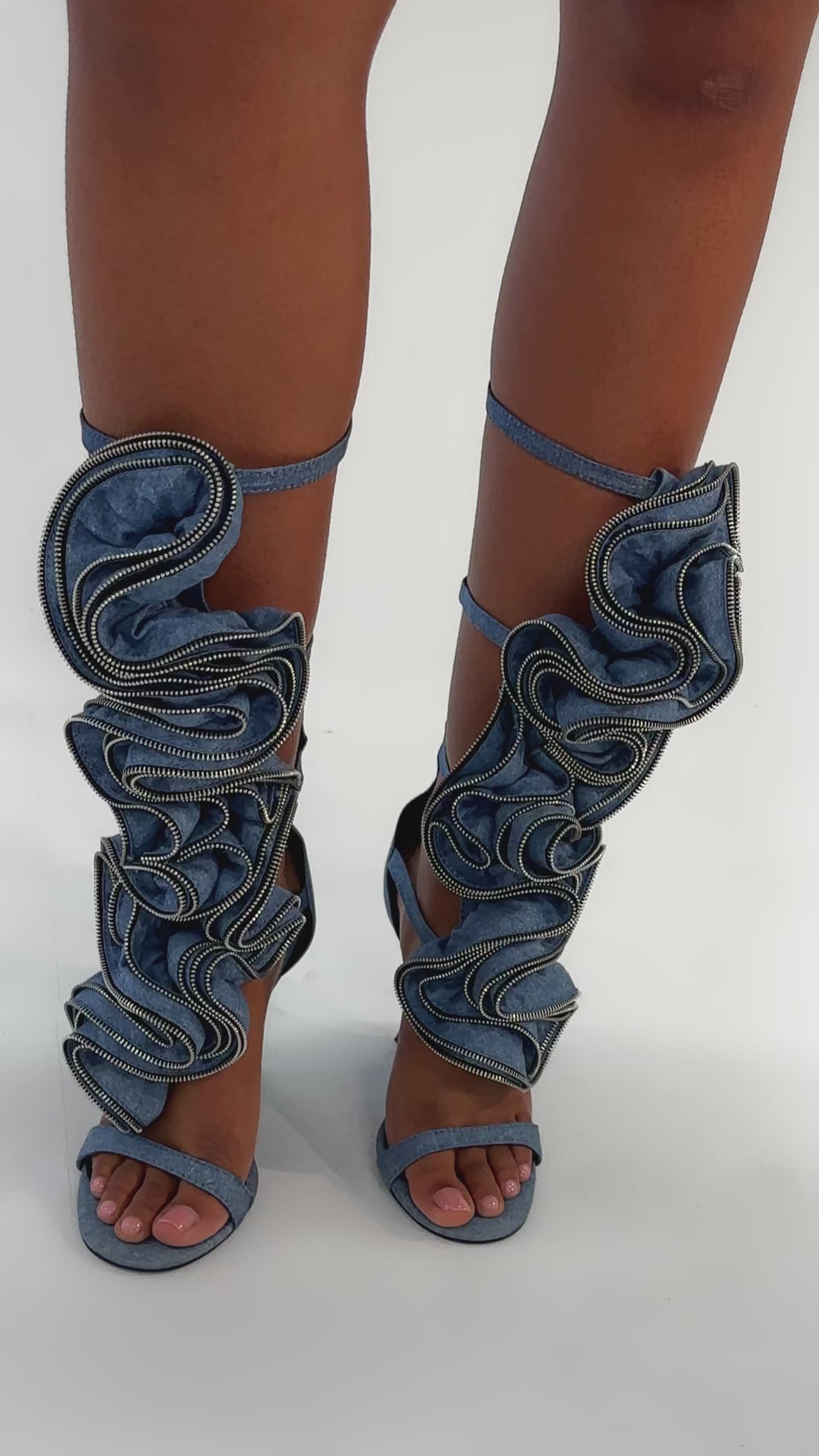 Video of woman modeling the AZALEA WANG ATLANTIC DENIM GLADIATOR WEDGE: knee-high, open-toe blue denim sandals with dramatic ruffled layers and visible stitching, posed against a plain light background.