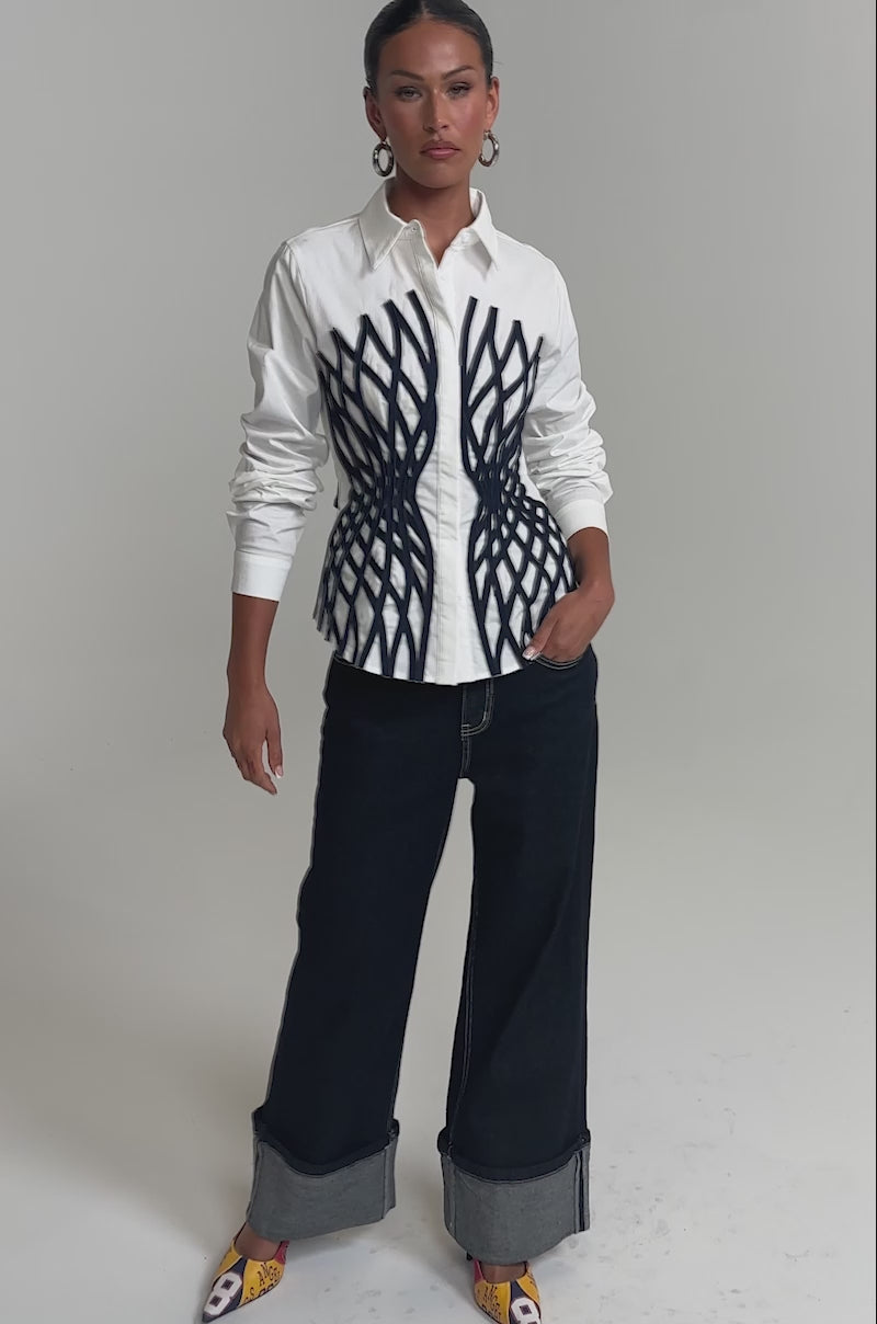 Video of woman modeling the ON A NEW LEVEL POPLIN BUTTONDOWN, a white blouse with a sculptural black geometric design, paired with dark jeans. Slicked-back hair and large hoop earrings add to her bold, modern style.