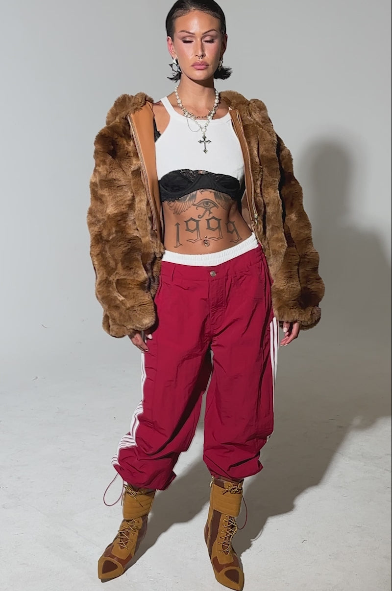 A video of a person in the LOOK AT ME NOW HOODED FUR BOMBER IN BROWN, a white crop top showing tattoos, red baggy track pants with white stripes, and tan lace-up boots against a plain background.