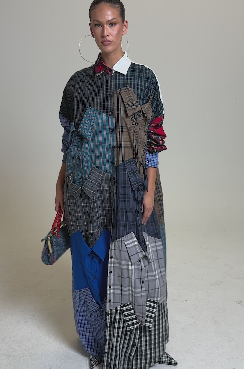 A video of a woman modeling the NOT YOUR BOYFRIENDS SHIRT DRESS, a long, oversized dress made of mismatched plaid shirt patterns. She carries a small blue patterned handbag and wears her hair slicked back against a plain gray background.