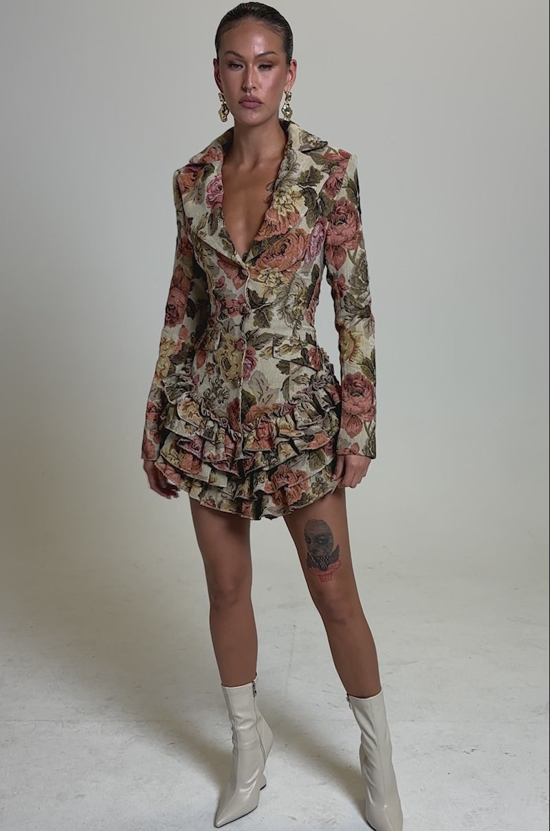A video of a woman posing confidently in the FLORAL HEAVEN TAPESTRY MINI DRESS featuring ruffled layers and a deep neckline, paired with light heeled boots, slicked-back hair, and gold hoop earrings.