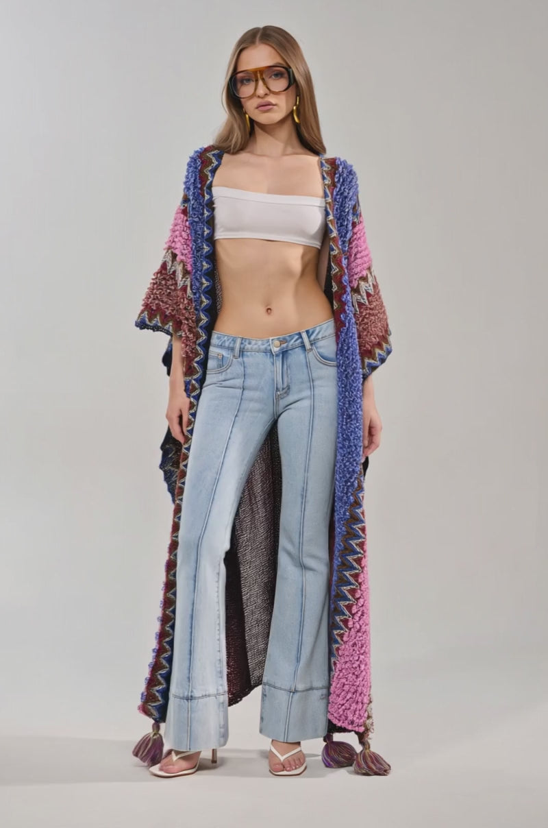 Video of woman modeling the WILDFLOWER WAVES LONG CARDIGAN—featuring bold blue and pink zigzag knits—layered over light blue jeans and silver heels, making it perfect festival wear against a simple background.