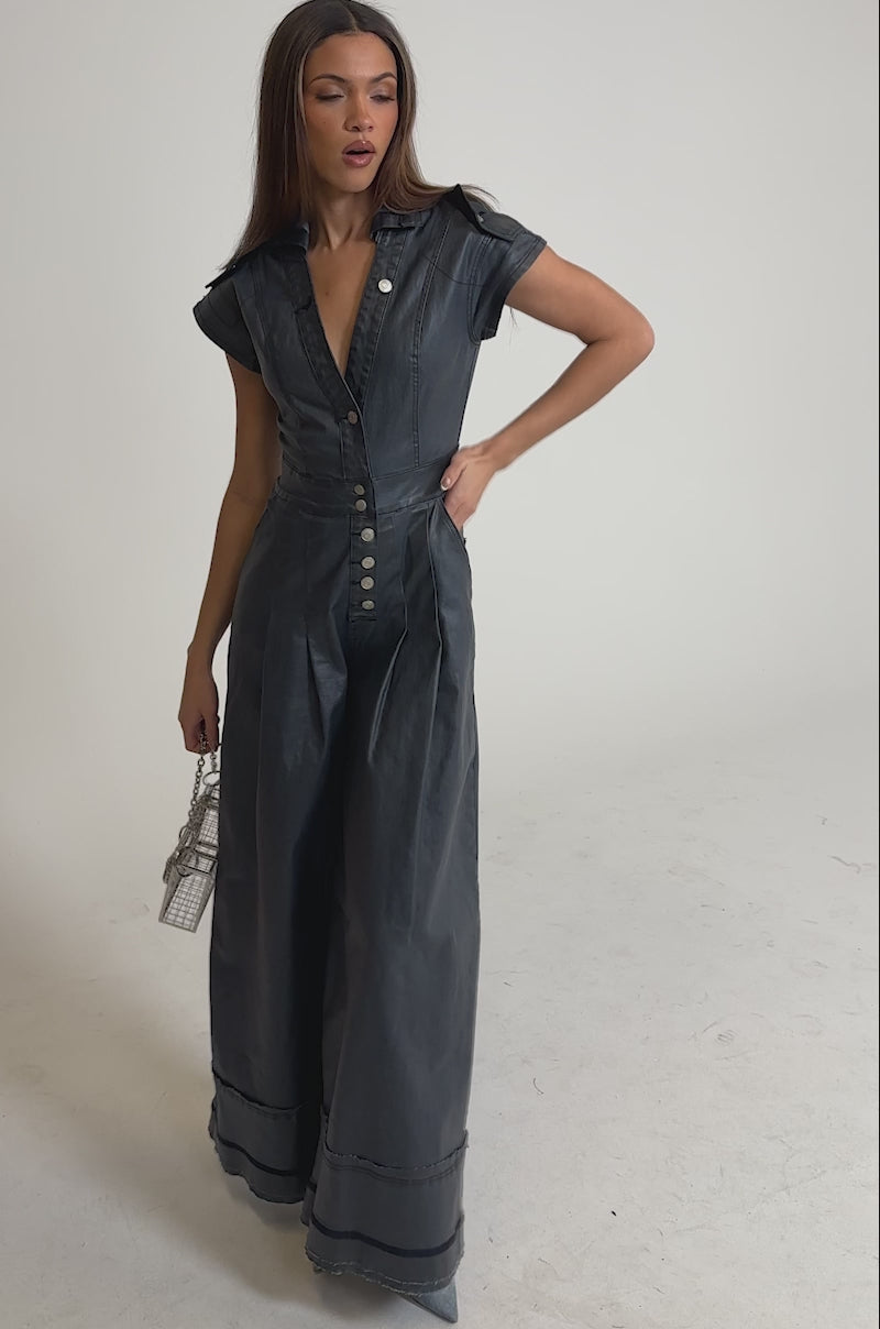 Video of woman modeling the RONNIE DENIM JUMPSUIT, featuring wide legs, a dark wash, short sleeves, and front buttons. She accessorizes with a chain-strap purse and light blue heels. Her long brown hair is straightened.