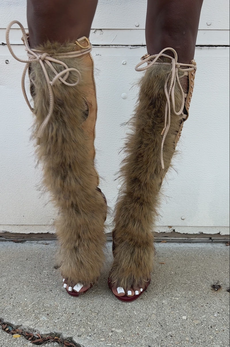 A video of a person standing on a sidewalk wearing the AZALEA WANG SHAINE MULTI GLADIATOR STITCH AND FUR SANDAL, featuring knee-high stiletto heels, faux fur accents, and open toes.