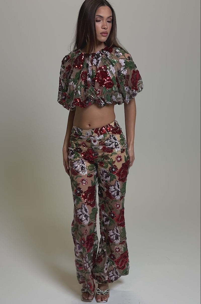 Video of woman modeling the ENCHANTED GARDEN FLORAL EMBROIDERED TULLE TOP, featuring puffed sleeves and vivid red, green, and purple floral patterns on sheer tulle, styled with matching high-waisted pants against a plain background