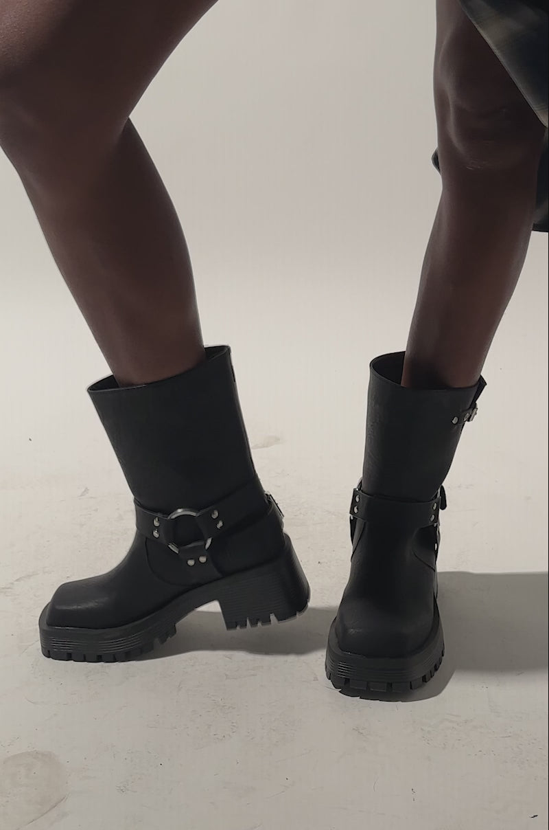 A video of a person wearing a plaid skirt and AZALEA WANG BUCKED BLACK CHUNKY HEEL COMBAT BOOTIE with silver buckles standing on a light background; only their legs and the lower part of the skirt are visible.