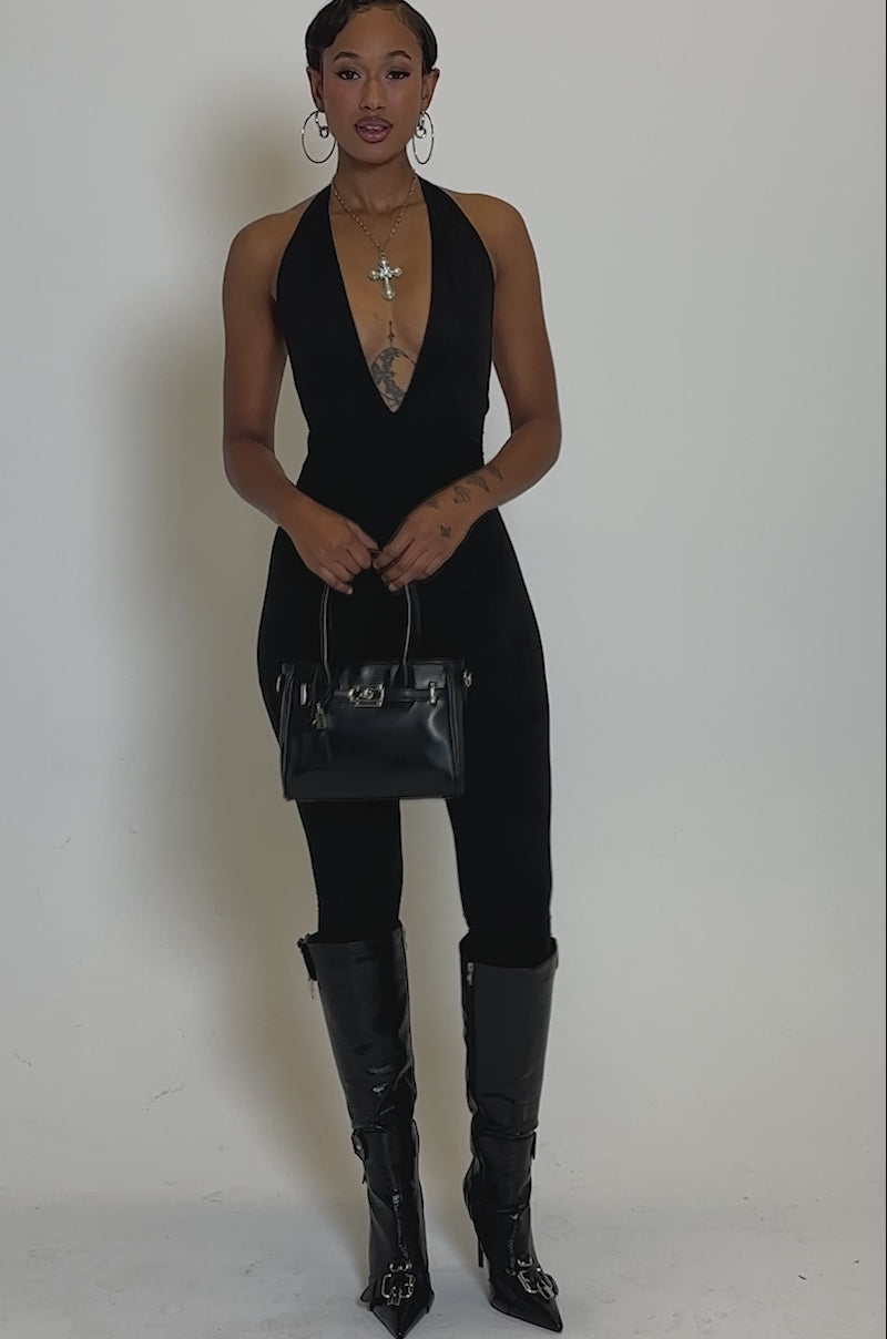 Video of woman modeling the GIA SUEDE JUMPSUIT, featuring a black, curve-hugging, halter style, with high heels, a black handbag, and hoop earrings against a plain light background.
