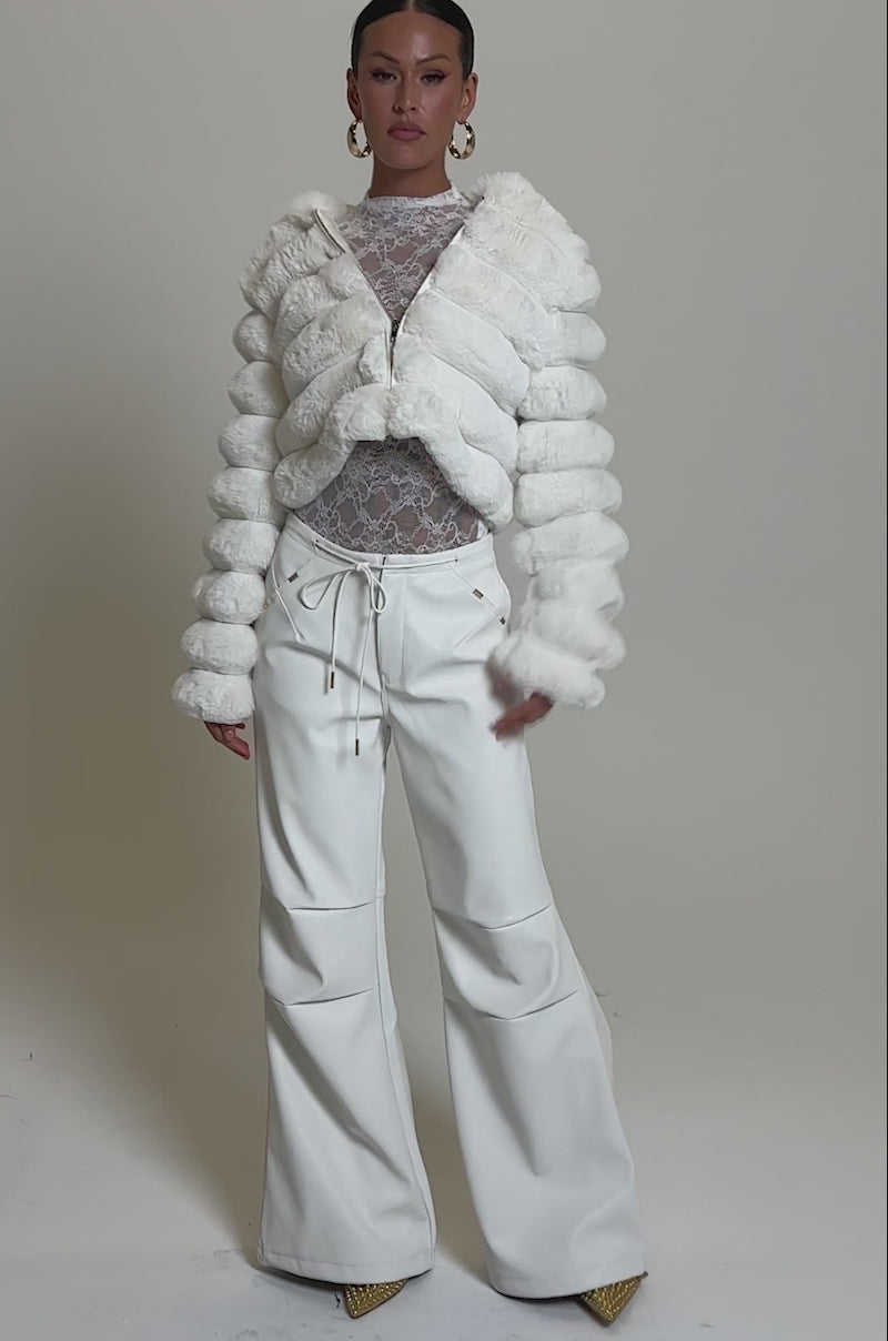 A video of a woman wearing the GET WITH IT FAUX LEATHER TROUSER IN WHITE, styled with a white fur jacket, sheer lace top, and gold heels.