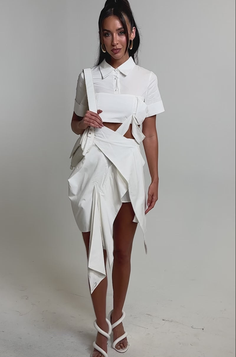 A video of a woman wearing the RIRI CUT OUT POPLIN MIDI DRESS, a modern white dress with cut-out detailing, paired with a white handbag and high-heeled sandals.