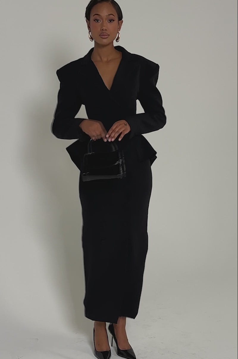 Video of woman modeling the KIMMY BLAZER MAXI DRESS, a long, fitted black suiting piece with double-breasted detail, paired with black heels and a small handbag against a light background.