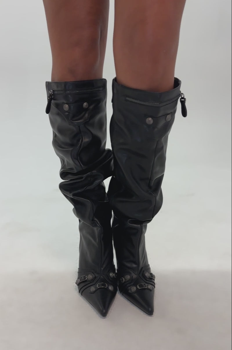 Video of woman modeling the AZALEA WANG TESARA POINTED TOE HARDWARE EMBELLISHED BOOT—black, slouchy, knee-high stiletto boots with straps and studs—standing against a plain gray background.