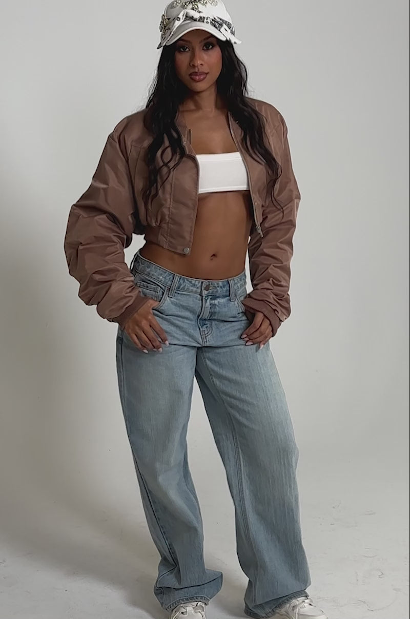 Video of woman modeling the TAKE IT EASY CROPPED BOMBER IN MAUVE over a white crop top, light blue jeans, and a white embellished baseball cap.