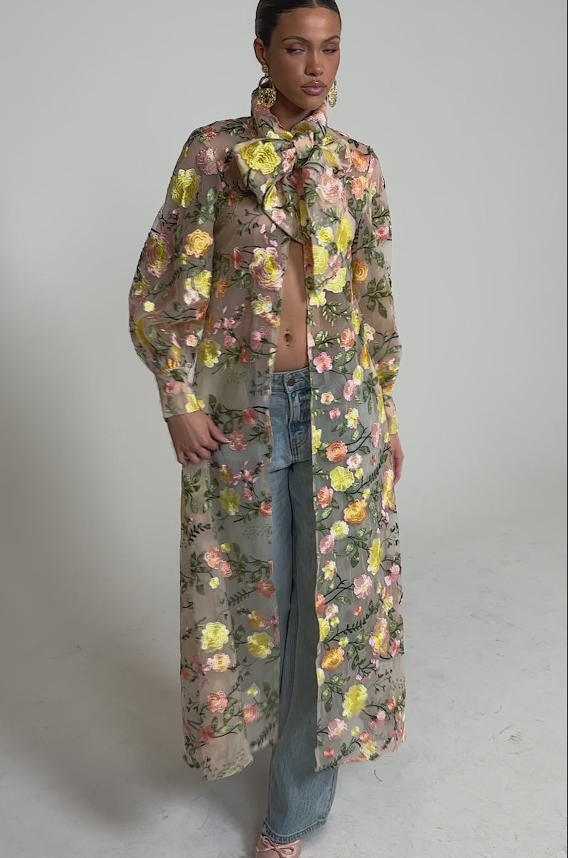 A video of a woman wearing the BOUQUET DREAMS FLORAL TULLE DUSTER—a sheer maxi coat with pastel pink, yellow, and green embroidered flowers and a statement neck bow.