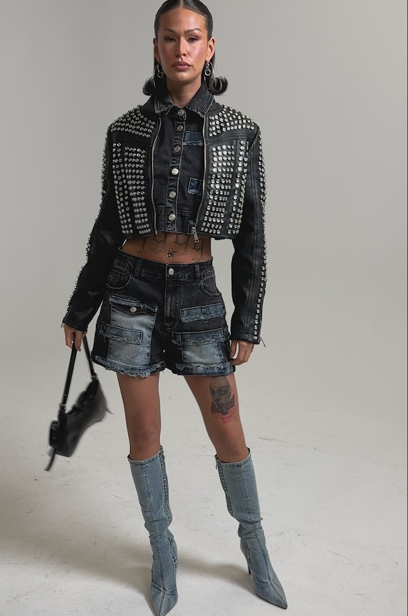 A video of a model wearing the ASHTON RHINESTONE MOTO jacket, dark patchwork denim shorts, blue knee-high boots, holding a small black handbag.