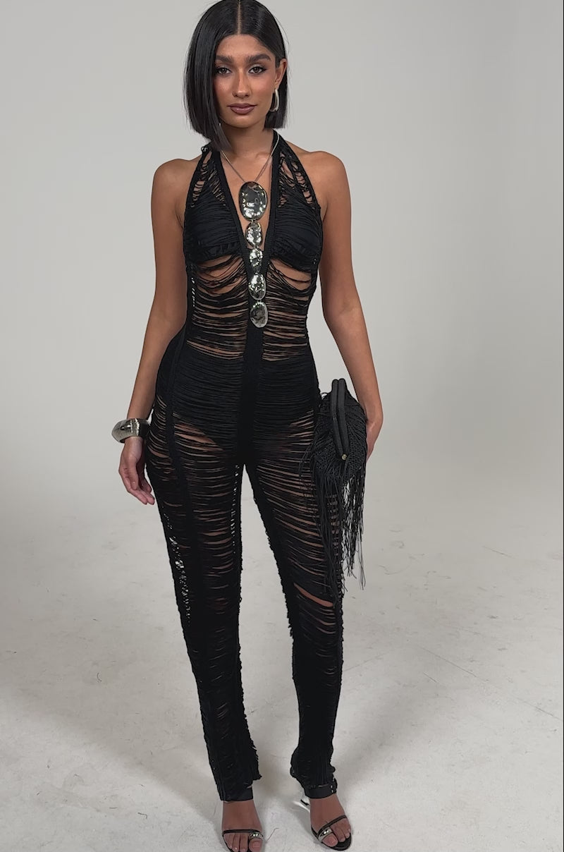 Video of woman modeling the IT GIRL SHREDDED JUMPSUIT, a woman stands confidently in this sheer black festival look with bold metallic front details, accessorized with a fringed black clutch, open-toe heels, and chunky bracelets.