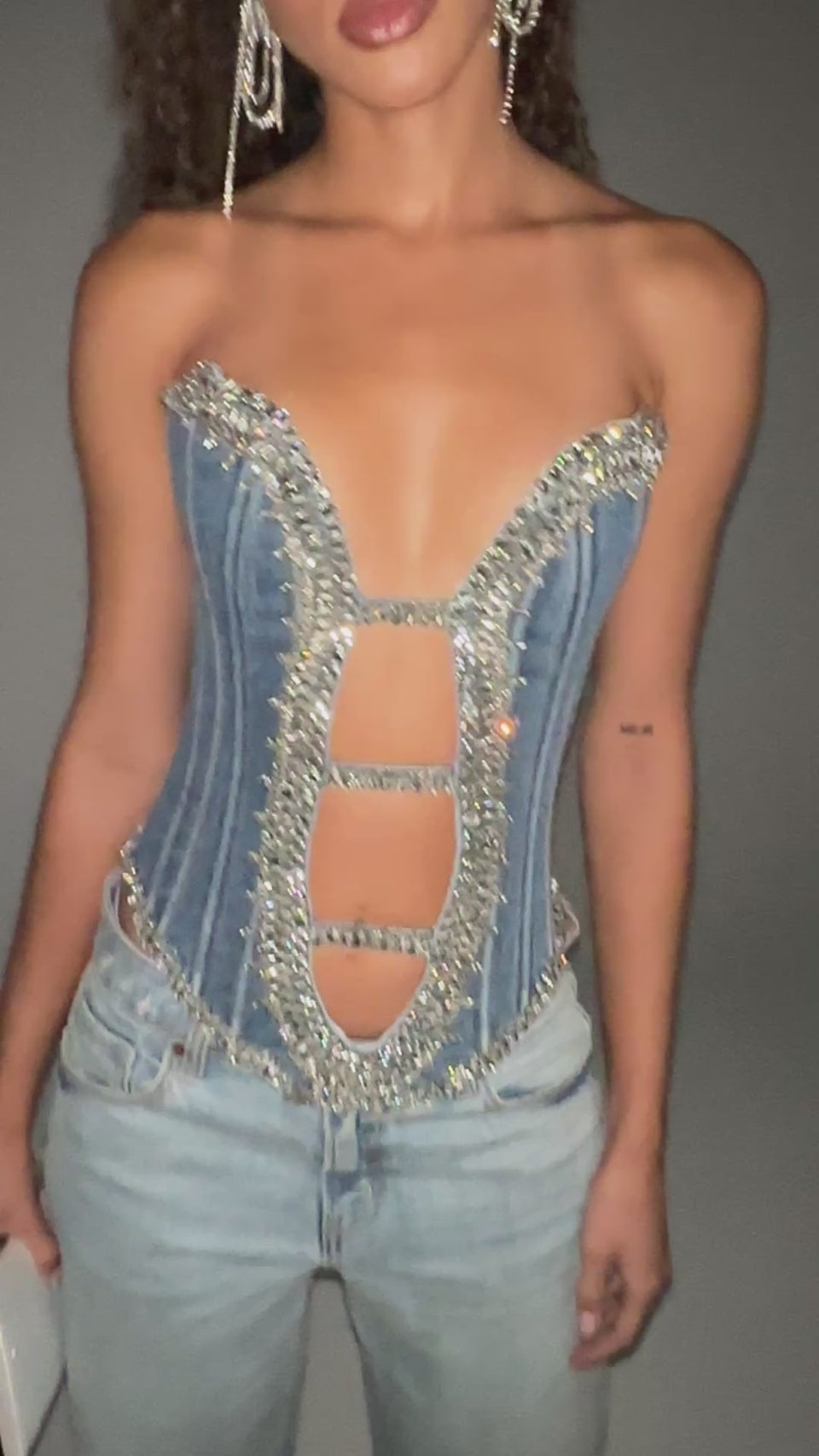 A video of a LUX LIFE CORSET IN LIGHT BLUE DENIM. Crafted from light wash stretch denim, this stunning corset is adorned with coruscant crystal embellishments that outline the bust, torso, and hem for a dazzling, opulent effect. It also features a strapless silhouette, cut-out ladder details at the center front, internal structural boning, smooth inner lining, and a back zipper closure.
