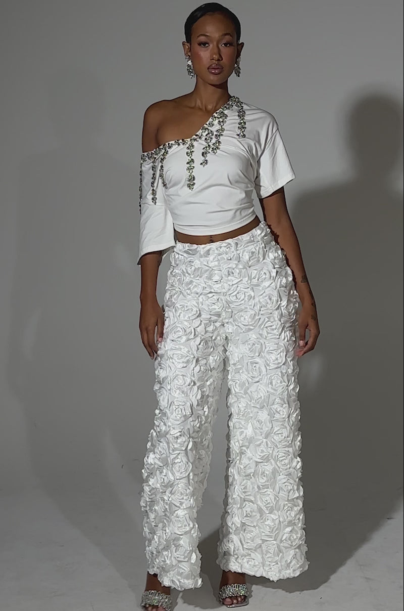 A video of a woman with short hair, hoop earrings, and strappy heels poses in the ROSE PETAL WIDE LEG PANT—white wide-leg pants adorned with satin rose appliques—and a white asymmetric top with silver details against a plain background.